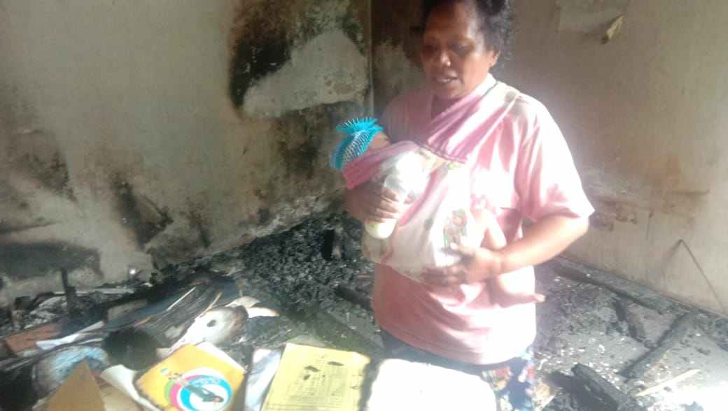Nona Elwuar sambil.menggendong anak melihat kondisi rumah orang tua yang terbakar dan rusak